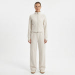 A model stands against a plain white studio background wearing a matching light beige zip-up jacket and wide-leg sweatpants set.