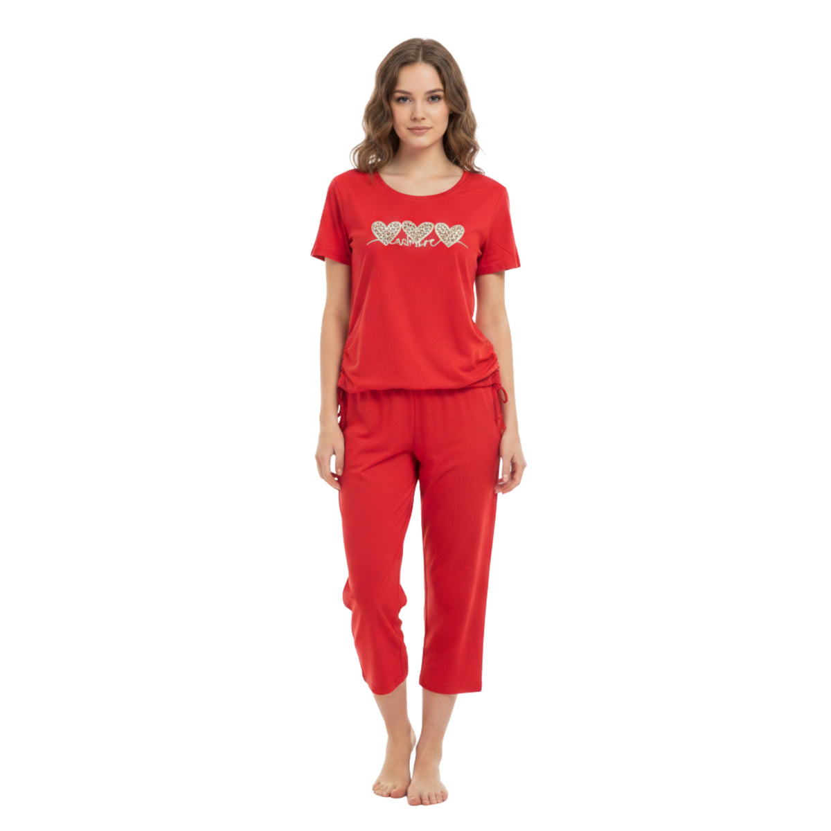 A woman modeling a red two-piece pajama set consisting of a short-sleeve t-shirt with a heart pattern on the chest and matching red cropped pants, shot against a plain white background.