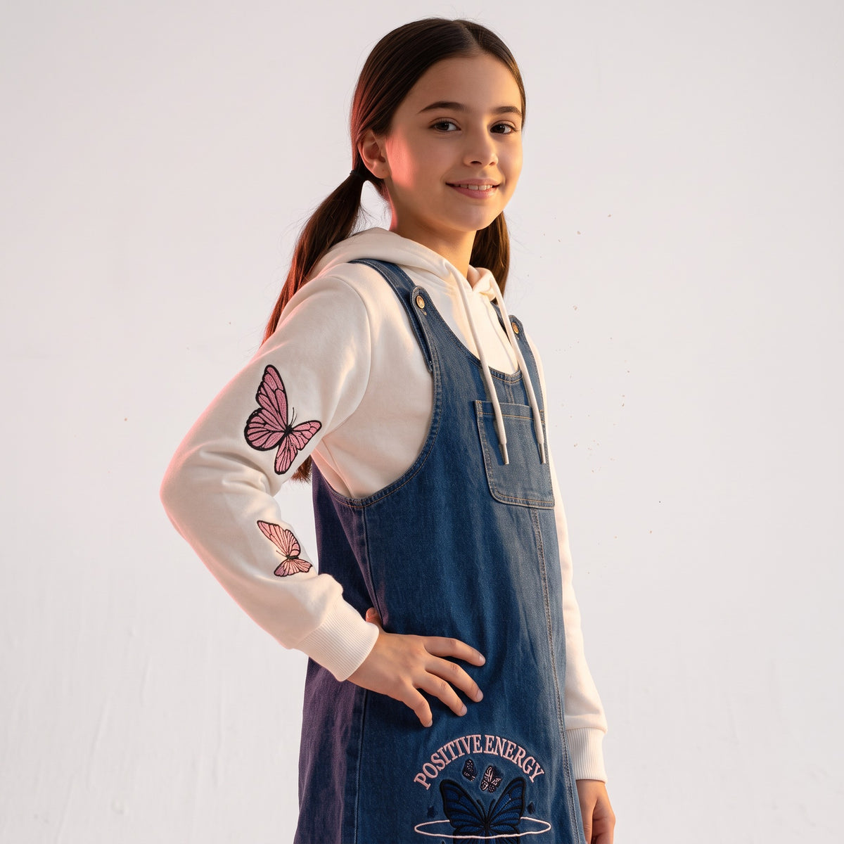 JOANNA FASHION girl wearing a blue denim pinafore dress over a white hoodie with pink butterfly sleeve graphics