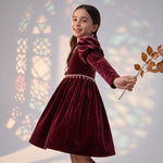A girl wearing a JOANNA FASHION deep red velvet dress with puff sleeves and pearl trim, holding a dried branch.