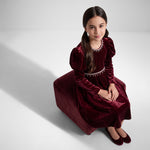 JOANNA FASHION girl in a deep red velvet dress with puff sleeves and pearl trim sitting on a matching velvet stool
