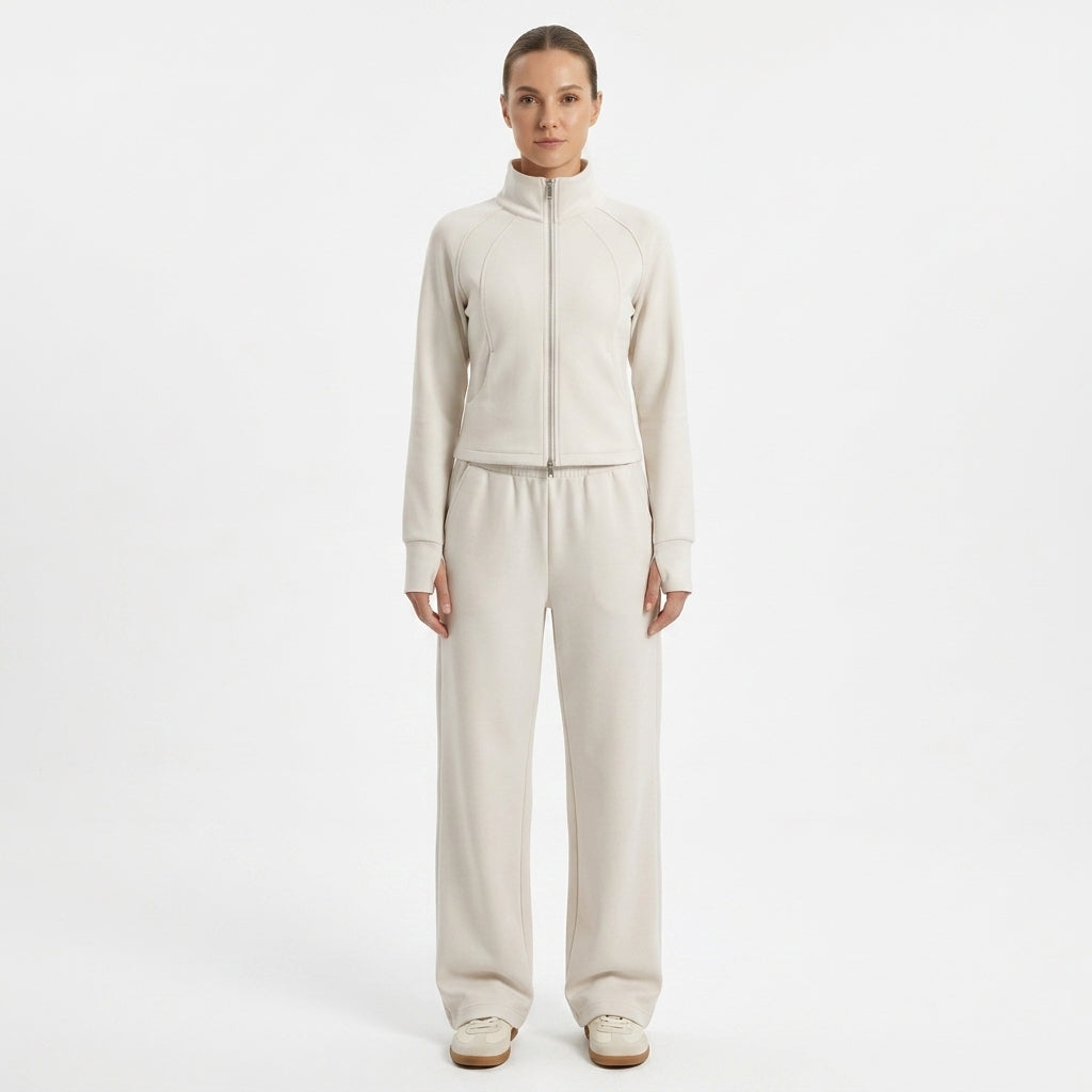 A model stands against a plain white studio background wearing a matching light beige zip-up jacket and wide-leg sweatpants set.
