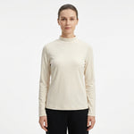 A woman modeling a long-sleeve mock neck knit top in a light cream color, paired with black pants, against a plain white studio background.