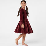 A girl wearing a JOANNA FASHION long-sleeve burgundy corduroy dress with white button details at the waist.
