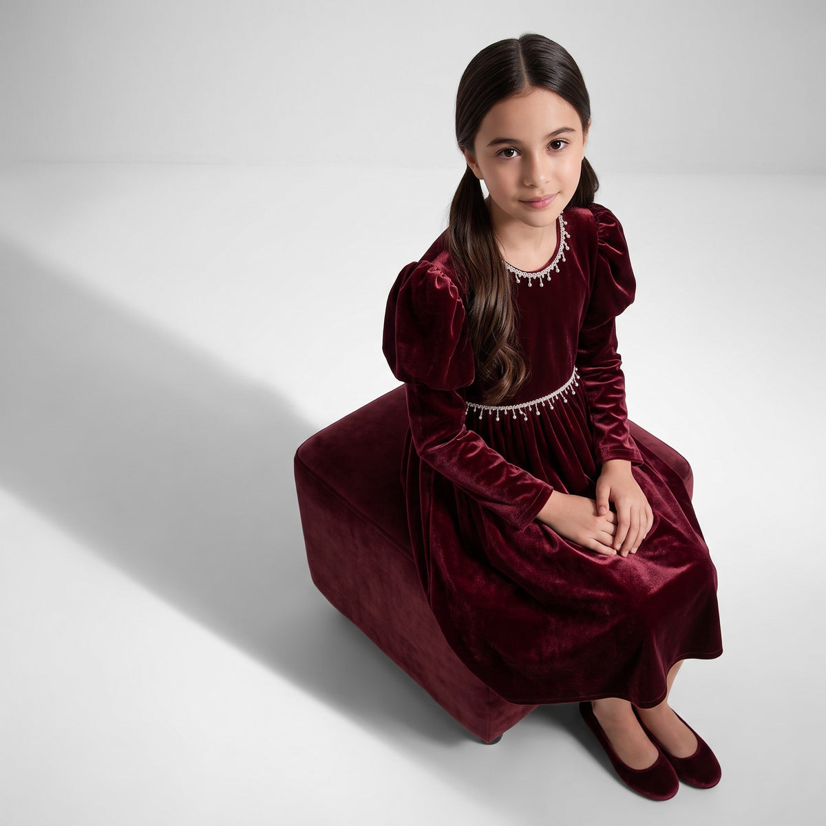 JOANNA FASHION girl in a deep red velvet dress with puff sleeves and pearl trim sitting on a matching velvet stool