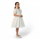 A young girl smiling in a white JOANNA FASHION formal dress with a lace bodice, puff sleeves, and a tulle skirt.