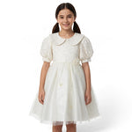 A young girl wearing a white JOANNA FASHION formal dress with a lace bodice, peter pan collar, and tulle skirt.